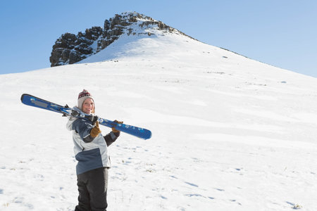 Woman carrying pair of alpine skis on shoulder wearing ski jacket on snow-covered slope, copy space. Adventure, winter sport, outdoor, extreme, athletic, wilderness, pristineの写真素材