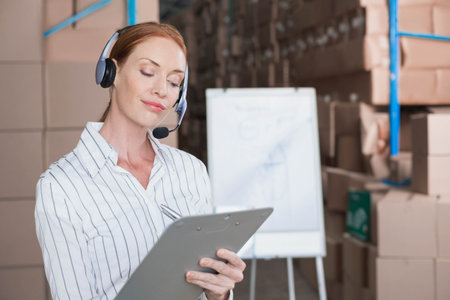 Woman standing in warehouse using headset writing clipboard notes near flip chart, copy space. Logistics, inventory, management, industrial, organization, productivity, teamworkの写真素材