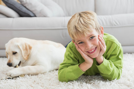Blond boy lying on white shag rug at home smiling while puppy chewing bone near sofa. Playful, childhood, relaxation, cozy, natural light, domestic, leisureの写真素材