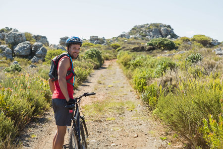 Male cyclist pausing beside mountain bike wearing helmet on dirt trail through rocky scrubland....