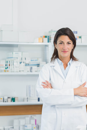 Female pharmacist in white lab coat standing arms crossed in pharmacy by pill bottles, copy space. Medical, professional, clinical, health, science, medication, consultationの写真素材