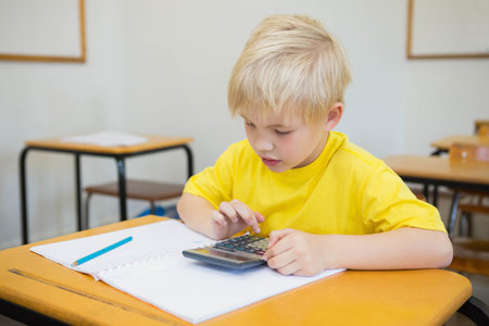 Boy in yellow T-shirt sitting at wooden desk in classroom using calculator and open notebook pencil....