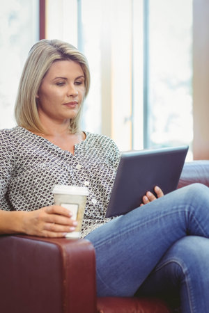 Woman sitting in leather armchair at home beside windows holding digital tablet and sipping coffee....