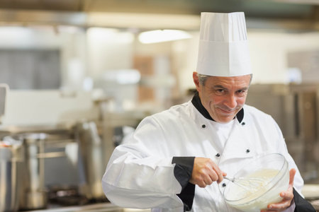 Male chef wearing toque whisking mixture with metal whisk in bowl in restaurant kitchen, copy space....