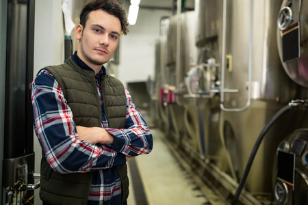 Male brewer wearing green vest and plaid shirt standing in brewery corridor by fermentation tanks....