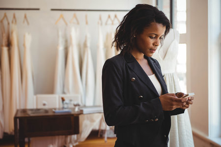 African American woman checking smartphone at bridal shop by table, mannequin and gowns, copy space. Fashion, retail, boutique, craftsmanship, elegance, style, professionalismの写真素材