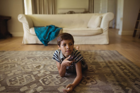 Boy lying on patterned rug in living room holding remote control and watching TV. Family room, relaxation, cozy, leisure, modern, interior, casualの写真素材