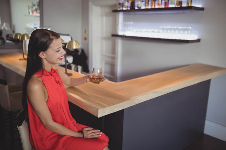Woman sipping drink from short glass at bar with backlit liquor bottles, copy space. Cocktail, interior, nightlife, elegance, hospitality, ambiance, loungeの写真素材