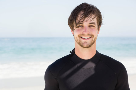Man standing near waterline on sandy beach wearing black wetsuit and smiling, copy space. Adventure,...