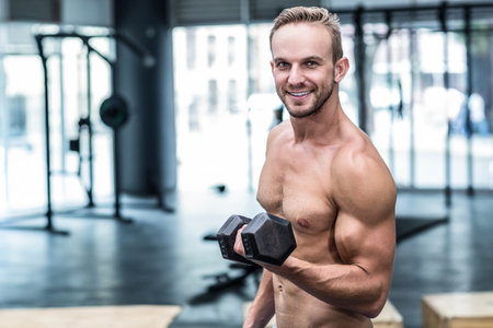 Shirtless mid-20s male lifting hexagonal dumbbell at waist in gym with squat rack and windows. Athletic, strength, training, fitness, minimalist, masculine, functionalの写真素材