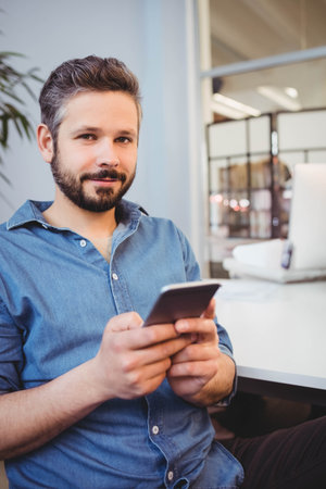 Man in denim shirt sitting at desk in office holding smartphone with laptop, glass partition, plant. Professional, coworking, modern, lifestyle, technology, workspace, corporateの写真素材