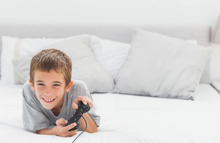 Boy lying on bed holding black game controller and smiling by pillows in bedroom, copy space. Youth, relaxation, entertainment, leisure, cozy, casual, modernの写真素材