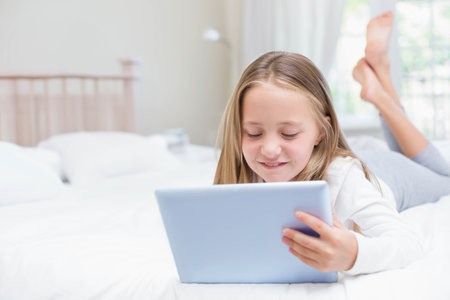 Schoolgirl lying on bed holding tablet under sheer curtains beside lamp in bedroom. Playful, cozy, serene, minimalist, digital learning, home interior, natural lightの写真素材