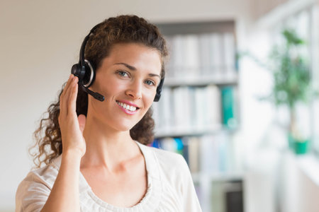 Woman adjusting headset earpiece while smiling in office near window, binder shelves and plant....