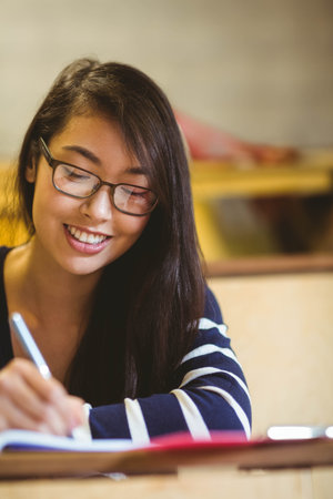 Asian woman sitting at desk in study room wearing glasses writing in notebook with silver pen. Educational, study, academic, library, concentration, casual, professionalの写真素材