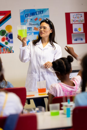 Asian teacher in a lab coat holds a beaker with a yellow liquid in a school classroom. She is...