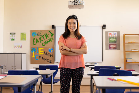 Asian teacher stands confidently in a school classroom. She is a teacher, with educational posters...
