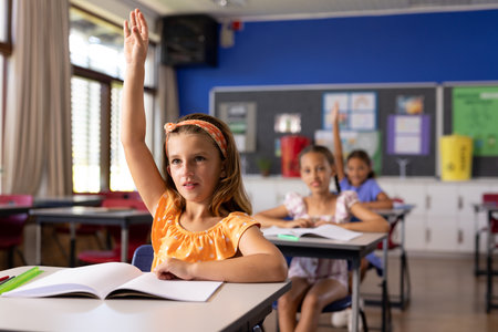 Caucasian girl raises her hand in a school classroom with copy space. Two girls sit behind her, engaging in a typical school setting with focus and curiosity.の写真素材