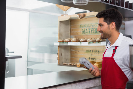 Male cashier wearing red apron offering card reader at bakery counter with bread shelf, copy space....