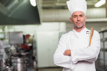 Senior male chef in chef uniform holding wooden spoon at shoulder in commercial kitchen, copy space....