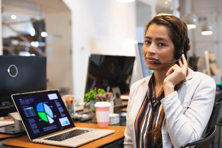 Woman in blazer handling calls with headset using laptop showing charts in office, copy space....