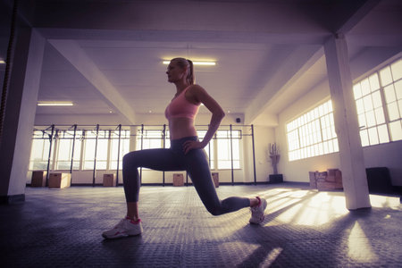 Woman performing lunge on textured rubber gym floor by pull-up rig, rope, plyo boxes near windows. Fitness, strength, training, modern, wellness, sport, motivationの写真素材