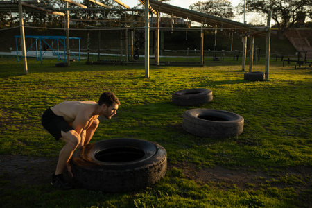 Man wearing black shorts squatting on field flipping tractor tire near obstacle frame, copy space....