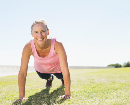 Mature adult woman holding plank on grassy field at park showing athletic shoes and leggings....