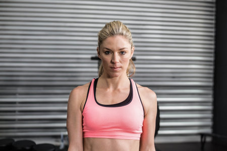 Woman in pink activewear, standing in gym before metal shutter with dumbbell rack and bench....