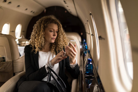 Businesswoman tapping smartphone screen in private jet cabin with blue water bottle. Luxury,...