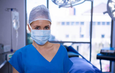Female surgeon wearing blue scrubs and mask preparing for surgery in surgical suite with IV monitor. Medical, healthcare, clinical, precision, sterile, emergency, professionalの写真素材