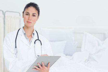 Female physician standing in hospital patient room holding clipboard with stethoscope by bed. Medical, healthcare, professional, clinical, diagnosis, hospital, wellnessの写真素材