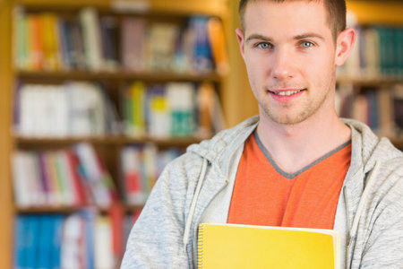 Male student standing before wooden bookshelves and holding yellow notebook at library, copy space....