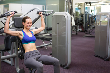 Female gym goer wearing sports bra and leggings using shoulder press machine at gym, copy space....