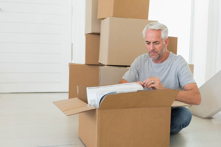 Man kneeling on floor in bright home unwrapping bubble-wrapped item from cardboard box, copy space....