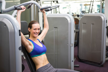 Adult woman wearing sports bra pushing chest press machine handles at gym by mirrors, copy space....