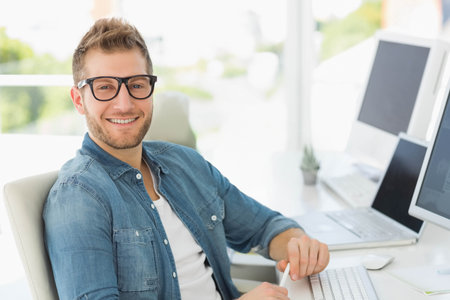 Man wearing denim shirt and glasses sitting at office desk typing on laptop by potted succulent....