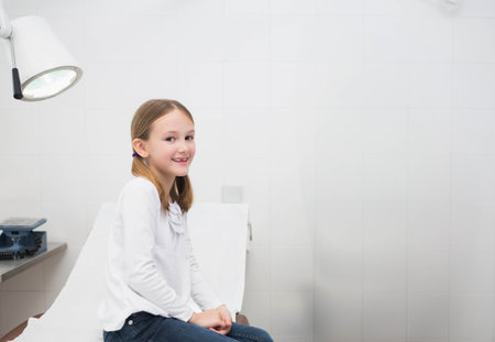 Preteen girl smiling and sitting on examination table in clinic with lamp and scale, copy space. Pediatric, healthcare, clinical, wellness, youth, medical, cleanlinessの写真素材