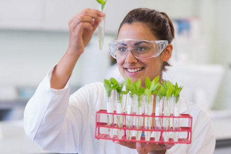 Woman wearing lab coat and goggles examining green seedlings in red test tube rack in laboratory. Science, research, innovation, biotechnology, growth, experimentation, developmentの写真素材