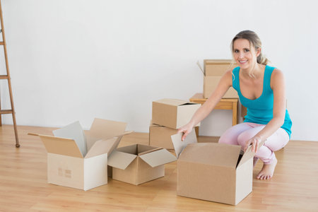 Woman kneeling unboxing cardboard boxes inside new home with ladder and desk, copy space....