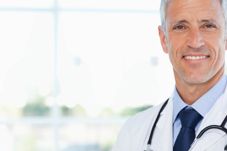 Senior male doctor standing in office by window wearing lab coat holding stethoscope, copy space. Clinical, healthcare, professionalism, consultation, wellness, diagnostics, natural lightの写真素材