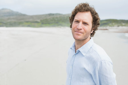 Middle-aged man standing on coastal beach overlooking distant hills and calm shoreline, copy space....