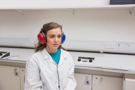 Woman in twenties sitting in exam room wearing lab coat, red and blue headphones using audiometer. Clinical, diagnostic, healthcare, diagnostic equipment, medical, technology, professionalの写真素材
