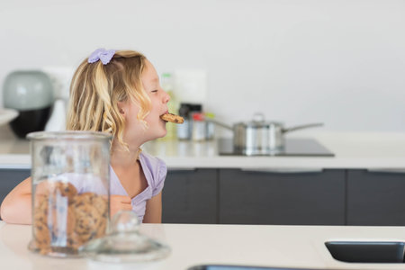 Child girl reaching for chocolate chip cookie from glass jar on white kitchen counter, copy space. Playful, cozy, modern, home, interior, innocence, snackの写真素材