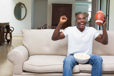 Mid adult African American man holding football and popcorn cheering on sofa at home, copy space. Celebration, leisure, entertainment, cozy, rustic, excitement, casualの写真素材
