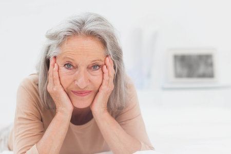 Senior woman lying on bed in bedroom, leaning on hands among books and focusing on laptop. Elegant, contemplative, serene, modern, cozy, lifestyle, natural lightの写真素材