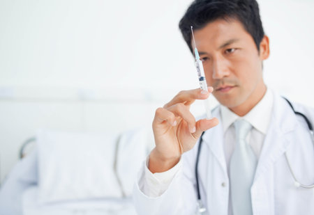 Asian male doctor inspecting syringe needle with stethoscope on neck at clinic bed, copy space. Medical, healthcare, professionalism, clinical, diagnostic, sterilization, safetyの写真素材