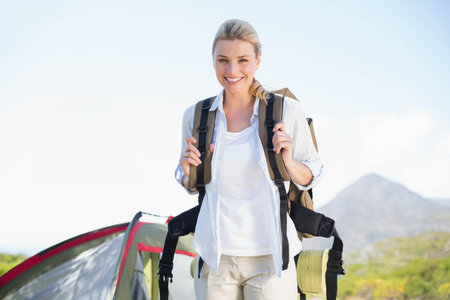 Female hiker standing in grassy clearing holding backpack with sleeping mat near camping tent....