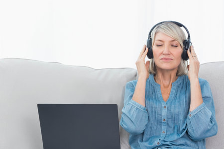 Senior woman sitting on sofa at home wearing denim shirt and headphones with laptop, copy space....