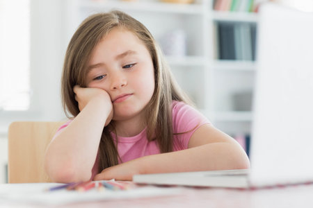 School-age girl wearing pink T-shirt sitting at home study desk with colored pencils and laptop. Children, study, education, creativity, workspace, relaxation, natural lightの写真素材